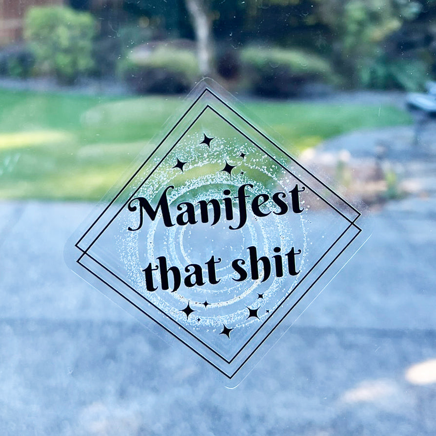 Vinyl decal stuck onto a window with the design double black squares with black stars and gold sprinkle dust black text that read Manifest that shit
