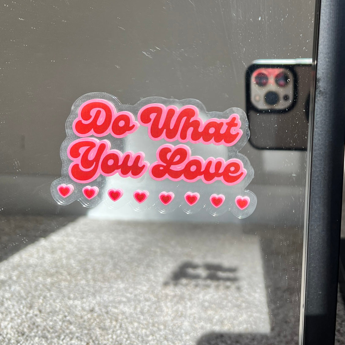 Window and mirror cling reusable sticker clinging to a mirror with phrase "Do What You Love" in red cursive font with pink outline and eight little hearts on the bottom