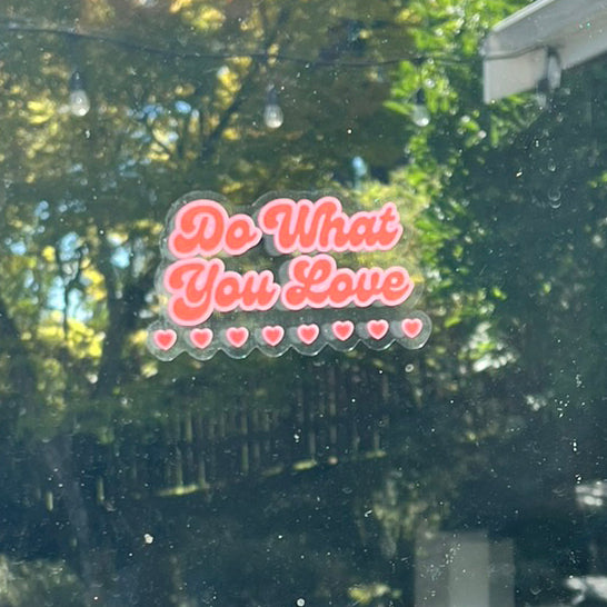 Window and mirror cling reusable sticker clinging to a window with phrase "Do What You Love" in red cursive font with pink outline and eight little hearts on the bottom